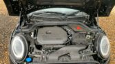 The image shows the engine compartment of a MINI Cooper with the hood open. The engine components and various parts are clearly visible, including the MINI-branded engine cover, air intake, coolant reservoir with a blue cap, and other components. The car is parked on a gravel surface.