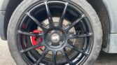 Close-up of a black alloy wheel with a multi-spoke design on a MINI. The wheel features a red brake caliper behind it, and there is tread visible on the tire. The wheel has a central hub cap bearing the brand logo "Bola." The background shows a portion of the car's grey body.