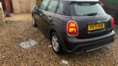 A dark-colored MINI Cooper with license plate PX71 XEB is parked on a gravel driveway in front of a wooden shed. The rear lights, designed in the shape of the Union Jack, add a distinctive touch. Surrounding the driveway is a wooden fence with green foliage visible in the background.