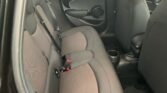 The image shows the backseat of a small MINI car with black and gray fabric upholstery. There are two rear seats with headrests, seat belts, and black interior door panels featuring circular design elements. The floor space includes a cup holder, and the back of the front seats is partially visible.