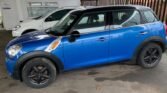 A blue Mini Cooper 1.6 with black rims is parked in a covered area beside a white car. The Cooper has black wheel arches and side trim. The ground is wet, and the background features a gray building with a window and a metal fence.