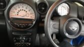 Interior of a COOPER 1.6, focusing on the dashboard and steering wheel. The speedometer is prominently centered, with a tachometer to the right. Below the speedometer are control buttons and displays. The steering wheel features the Mini logo. The overall color scheme is black.