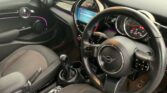 Interior view of a modern car featuring a sleek black dashboard with neon purple accent lighting. The MINI steering wheel, adorned with the Mini logo, has various control buttons. The center console includes a gear stick, multiple control knobs, and a digital infotainment display.