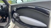 Close-up view of the interior passenger door of a MINI car. The door features a sleek black design with a metallic handle, circular air vent, speaker, and modern accents. The window is slightly open, and the exterior glass and greenery are visible in the background.