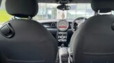 View from the backseat of a MINI, showing two front seats with black upholstery. The dashboard features a large circular speedometer in the center with red markings, and the steering wheel on the right side. The center console has controls and a gear shift. Outside, a building with large windows is visible.