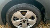 A close-up photo of a car wheel featuring a silver alloy rim with five spokes and a visible black tire on a MINI. The brand name "Sumitomo" is embossed on the tire's sidewall. The wheel is on a car parked on ground covered with small, multicolored gravel stones. The tire appears to be wet.