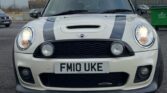 A white MINI Cooper with black racing stripes on the hood and roof is parked on an asphalt surface. The car boasts a front license plate reading "FM10 UKE." In the background, an overcast sky forms a subdued backdrop alongside a metal fence and some green containers.