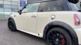 A white MINI Cooper with black trim and red brake calipers is parked in front of an industrial building with large glass windows. The car features black alloy wheels, a visible fuel cap, and a rear spoiler. A blue sign on the building reads "11 Orgreave Place.