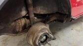 Close-up of a MINI's rear wheel area with the wheel removed, exposing a rusted brake drum and suspension components. The car's exterior is red, and part of the wheel well liner is visible. The ground beneath the vehicle is dirty, with what appears to be minor debris scattered around.