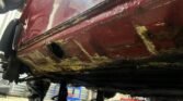 Close-up view of the undercarriage of a red MINI, showing significant rust damage. The car is elevated on a jack, revealing rust and corrosion along the bottom edge and frame. In the background, a person in work attire stands in a workshop setting filled with tools and equipment.