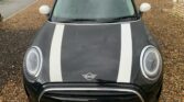 A black MINI Cooper with two white racing stripes on the hood is parked on a wet gravel driveway next to a beige brick building. The car has a white roof and white side mirrors, and its license plate reads "PX71 XEB." The pavement and surroundings appear damp from rain.