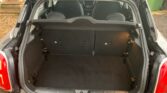 Image of the open trunk of a small black MINI, showing an empty cargo space with a dark carpeted floor. The back seats are upright and visible. The trunk area is clean and well-maintained, with the car’s exterior partially visible, glistening from recent rain.