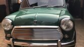 A classic Mini Cooper in green is parked on a brick driveway. The car features a prominent chrome grille, round headlights, and a chrome bumper with black rubber guards. The license plate reads "URM 527H." Inside the car, the steering wheel and part of the dashboard are visible through the windshield.