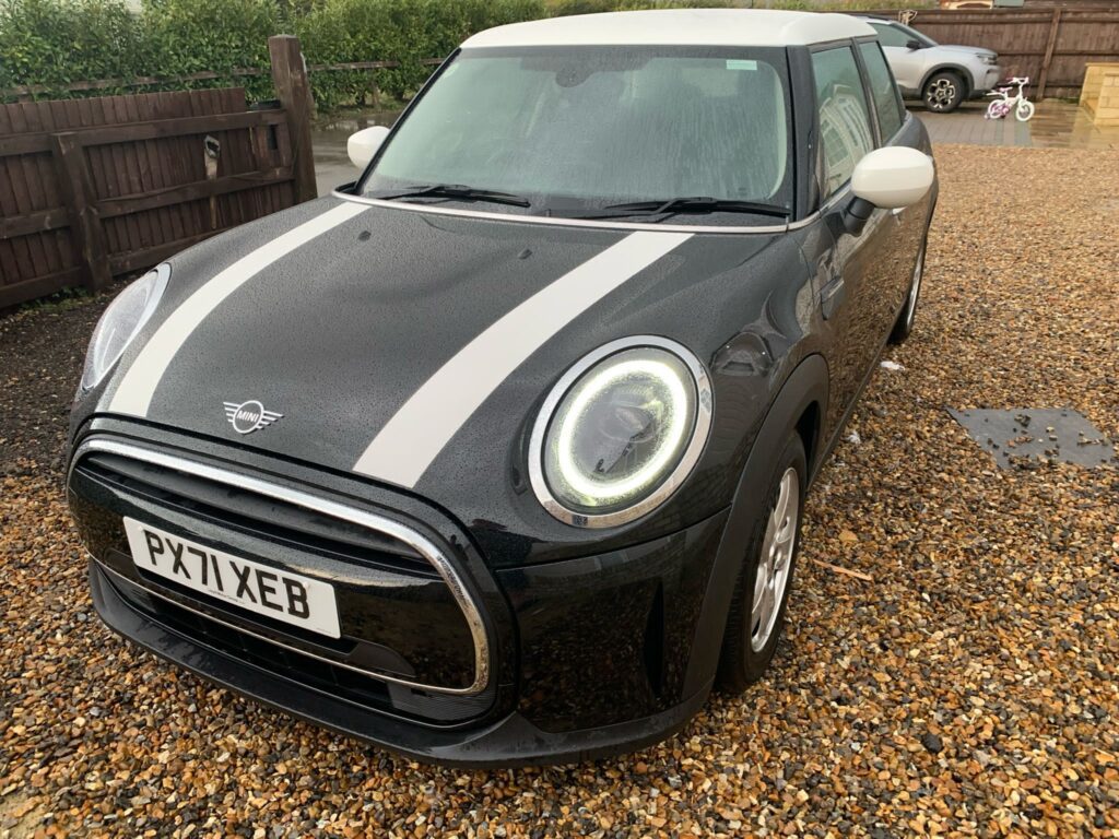 A black MINI Cooper with white racing stripes on its hood and a white roof is parked on a gravel driveway. The front license plate reads "PX71 XEB." The car's headlights are on, and there is a wooden fence and greenery in the background. A child's tricycle is also visible in the background.