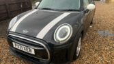 A black MINI Cooper with white racing stripes on its hood and a white roof is parked on a gravel driveway. The front license plate reads "PX71 XEB." The car's headlights are on, and there is a wooden fence and greenery in the background. A child's tricycle is also visible in the background.