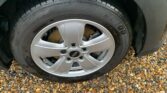 Close-up of a MINI's front wheel on a gravel surface. The wheel features a silver alloy rim with a five-spoke design and a black tire labeled "Hankook Kinergy GT" in raised letters. The car exterior around the wheel is black and appears to be wet from rain.