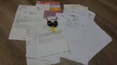 A collection of various documents laid out on a wooden floor, including sheets of paper, letters, and an envelope. Two black car keys with yellow tags, labeled MINI, rest on top of the papers. Among them is a colorful card, possibly a license, prominently visible among the papers.