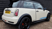 A cream-colored MINI Cooper S with a black roof is parked. The car features black alloy wheels, red brake calipers, and blue checkered decals reading "GP2 Supercharger" on the side. The rear license plate reads "LX06 UGT." The vehicle is positioned on a concrete surface against a dark wooden wall.