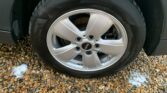 A close-up view of a car tire with a silver alloy wheel, featuring the MINI logo in the center. The tire is positioned on a bed of small, multi-colored gravel stones. The tread of the tire is visible, and a small amount of white foam is seen near the bottom left of the tire.