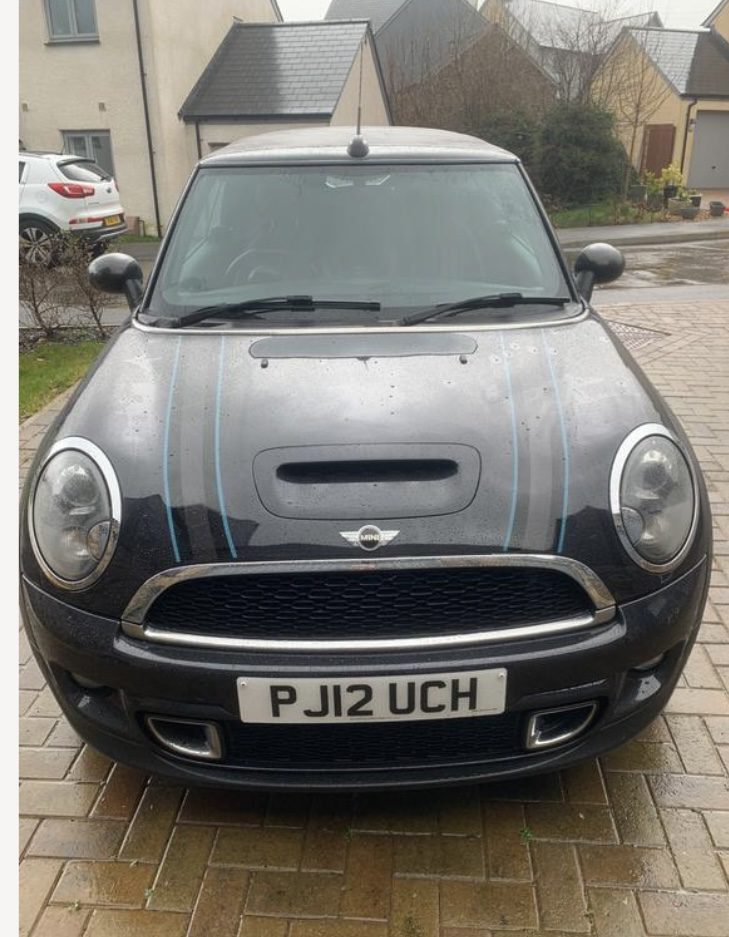 A black MINI Cooper with blue racing stripes is parked on a wet, brick driveway in front of suburban houses. The car has a British number plate with the registration "PJ12 UCH." Drops of water are visible on the MINI, indicating recent rain. Trees and a grey sky are in the background.