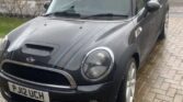 A black MINI Cooper with a gray racing stripe is parked on a wet driveway in front of a house. The car bears a UK license plate reading "P J12 UCH." Surrounding the scene are additional houses and a garden lush with greenery, all under the spell of rainy weather.