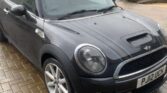 A gray MINI Cooper is parked on a brick driveway in front of beige-colored houses. Featuring sporty detailing with blue racing stripes on its hood and roof, the car stands out in the residential neighborhood on a cloudy day. The license plate reads "PJ12 UCH.