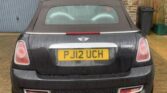 A black MINI Cooper S with a yellow license plate marked "PJ12 UCH" is parked on a brick driveway. The car's surface appears mildly dirty. Behind the vehicle, there are closed grey garage doors and a section of stone wall showcasing traditional brickwork. A green bin is visible to the right.