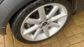 Close-up of a MINI's rear tire and wheel on a wet, brick-paved driveway. The wheel has a silver alloy rim with a five-spoke design. The tire and fender are wet, likely due to rain, evident from the water droplets. A partial reflection of a person is visible on the car's body.