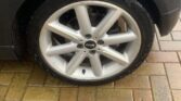 Close-up of a wet car tire and alloy wheel mounted on a dark grey vehicle. The vehicle, resembling a sleek MINI model, is parked on a wet brick-paved surface. The nine-spoke alloy wheel has a central hub displaying the BMW logo, with water droplets visible on both the tire and the vehicle.