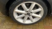Close-up of a wet car tire mounted on a silver, multi-spoke alloy wheel of a metallic dark gray MINI. The car is parked on a wet brick pavement. The tire is clean and shows no visible damage, with droplets of rain dispersed over it and the surrounding surface.
