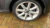 A close-up view shows the front left wheel of a dark-colored MINI Cooper parked on a wet, brick-pattern driveway. The car appears clean, and the tire is in good condition, with the MINI logo visible in the center of the alloy rim. The bricks vary in hue from light to dark brown.