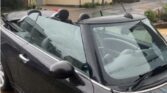 A sleek black MINI convertible with its top down is parked on a driveway, glistening from recent rain. In the background, several modern two-story houses with light and dark exteriors line a quiet residential street. The scene looks overcast.