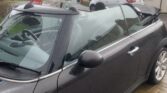 A black MINI convertible is parked on a wet brick driveway in a residential neighborhood. The car's retractable roof is down, exposing the interior. The weather appears to be rainy, as the MINI and ground are wet. Modern houses and other parked cars are visible in the background.