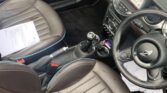 The image shows the interior of a right-hand drive MINI car featuring two brown leather seats, a steering wheel with the iconic MINI logo, and a speedometer in the center of the dashboard. There is a white sheet of paper on the passenger seat and various controls and buttons on the center console.