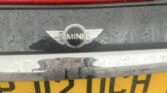Close-up of the rear of a black MINI car labeled with a "Mini" emblem above its license plate. The yellow plate reads "PJ12 UCH" and is slightly shiny from raindrops. Water droplets cover parts of the car's surface, creating a wet appearance.