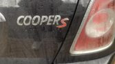 Close-up of the rear section of a black MINI Cooper S car, with water droplets on the surface suggesting recent rain. The "COOPER S" badge is visible on the trunk, and part of the right rear taillight is shown. A reflection of a person taking the picture is visible on the wet car surface.