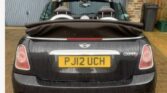 A black MINI Cooper convertible is parked in front of a two-door garage with stonework and a slate roof. The convertible top is down, exposing the rear seats. The car's license plate reads "PJ12 UCH," and there are two exhaust pipes at the back. The brick-paved driveway complements the sleek appearance.