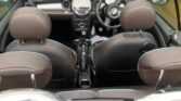 A view from above and behind of a black MINI convertible with its top down, showcasing the interior. The front seats are visible along with the steering wheel, dashboard, and gear shift. The car is parked in front of closed, light blue garage doors with a brick wall partially visible.