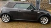 A black MINI Cooper convertible is parked on a brick driveway in front of a modern house with black and stone walls. The car's roof is up, and it has silver alloy wheels. The background includes several houses on a wet, overcast day.