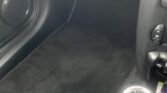 A car's interior focused on the front passenger side floor area. The black carpeted floor mat is clean and neatly placed. The edge of a brown dashboard and the gear shifter with a black leather boot are visible, revealing a MINI's six-speed manual transmission pattern.