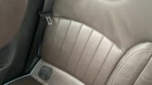 A close-up view of a brown leather MINI car backseat with blue piping. The seat has vertical stitching, two headrests, seat belt buckles, and two red seatbelt clips visible near the bottom right. The lighting suggests it was taken during the day.