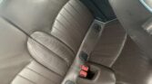 Close-up of a leather car seat with a ribbed design and white stitching in a MINI vehicle. The seat contains three black seatbelt buckles, one with a red release button visible. The image displays part of the seat and edges of the backrest, showcasing the texture and design details of the upholstery.