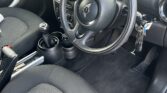 Interior of a MINI featuring a black steering wheel with various controls and a key inserted in the ignition. The dashboard has a large central speedometer. The front seats are dark gray fabric. The center console includes a gear shift, cup holders, and a small compartment. The door has a curved handle.