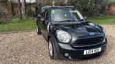 A black MINI Cooper is parked on a gravel driveway. The car, sporting the registration plate "LG14 BDE," is situated in front of a brick wall and tall trees. The outdoor scene features patches of green grass visible around the driveway. The MINI appears clean and well-maintained.