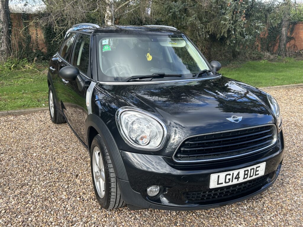 A black MINI Cooper with a white roof is parked on a gravel driveway. The vehicle's headlights and front grille are prominently visible. The license plate reads "LG14 BDE." Behind the car, there is a garden with green grass, trees, and a brick wall.