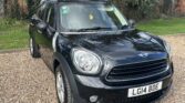 A black MINI Cooper with a white roof is parked on a gravel driveway. The vehicle's headlights and front grille are prominently visible. The license plate reads "LG14 BDE." Behind the car, there is a garden with green grass, trees, and a brick wall.