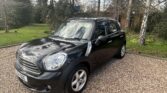 A black MINI Cooper is parked on a gravel driveway. The car is shiny and well-kept, with a four-door design and silver alloy wheels. It is surrounded by a lush green lawn, tall trees, and a wooden fence, suggesting a peaceful and secluded environment under a partially cloudy sky.