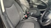 The image shows the interior of a black MINI, specifically the front seats. Both seats are upholstered in black fabric and have headrests. The car door on the driver's side is open, revealing the black interior and window control buttons. A parked car and greenery are visible outside.