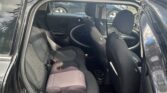 Interior of a black MINI photographed from an open passenger side rear door, showing two black and charcoal grey cloth seats with a child booster seat on the left. Trees and a McDonald's sign are visible in the background through the windows.