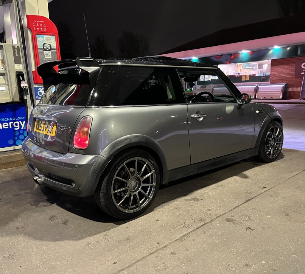 A grey MINI Cooper with black-tinted windows and custom black rims is parked at a gas station. The station is lit with overhead lights, and LED signs can be seen in the background. It's nighttime, evident by the dark sky and artificial lighting. The license plate reads "YA54 JFA".